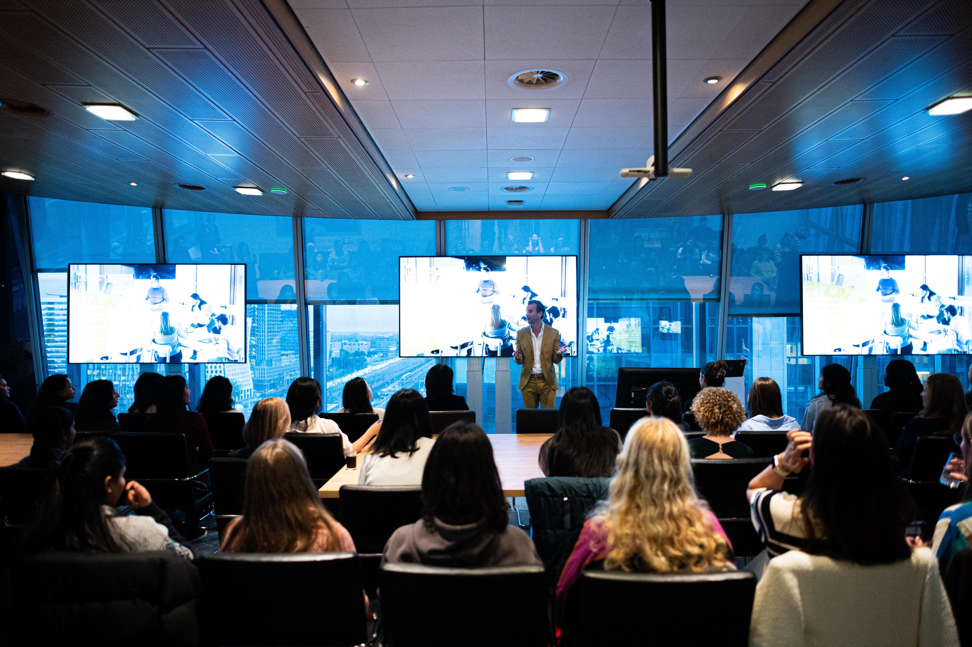 Uitzicht op de Zonnezaal, gevuld met aanwezigen die zitten en aandachtig luisteren naar een presentator vooraan die een presentatie geeft.